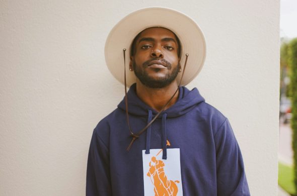Portrait of a fashionable man in a hat and hoodie standing against a light background.